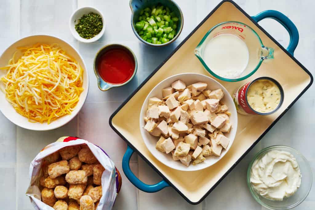 Overhead view of ingredients for buffalo chicken tater tot casserole: chicken, milk, soup, tater tots, cheese, celery, spices, sour cream and wing sauce.