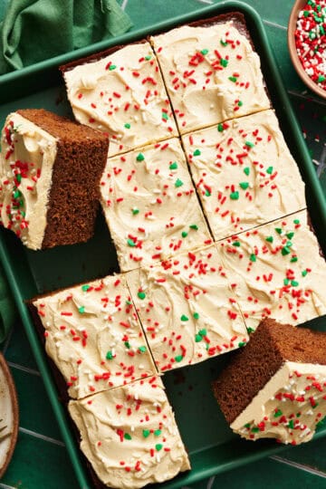 Gingerbread Cake with Cinnamon Molasses Frosting on a green sheet pan with a green napkin and tile background. Several slices removed from full cake.