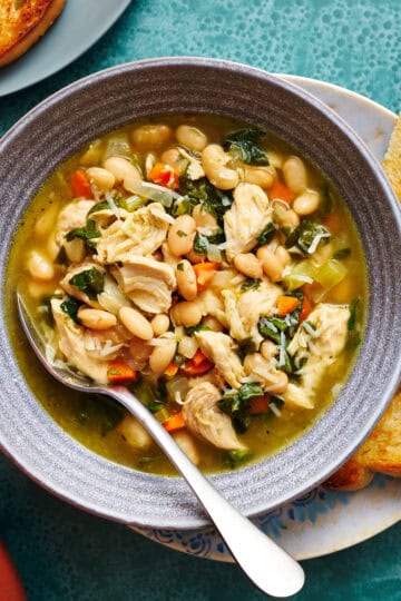 overhead view of a bowl filled with soup that has shredded turkey, beans, greens, broth, carrots and spinach. It has more bowls of soup surrounding and slices of bread on the side.