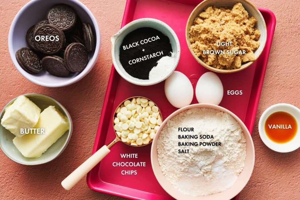 Overhead view of ingredients for Chocolate Cookies and Cream Cookies on a pink sheet pan. Includes Oreo cookies, black cocoa powder, cornstarch, light brown sugar, eggs, flour, baking soda, baking powder, salt, vanilla, butter and white chocolate chips.