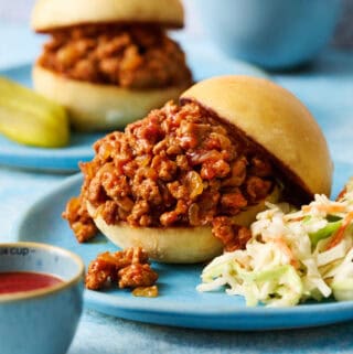 A ground chicken sloppy joes sandwich on a blue plate with coleslaw with another sandwich in the background and cheese in a bowl.