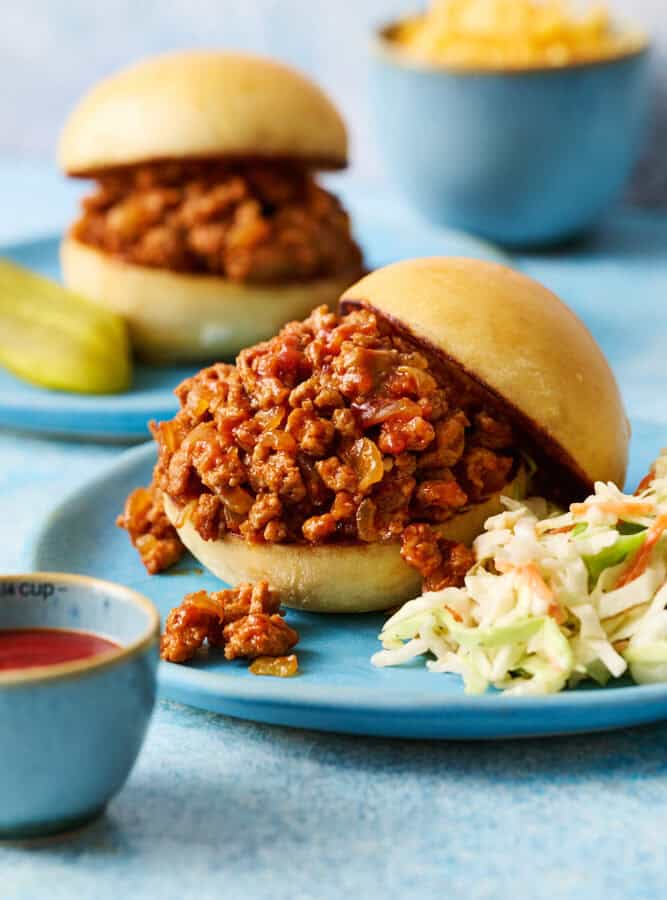 A ground chicken sloppy joes sandwich on a blue plate with coleslaw with another sandwich in the background and cheese in a bowl.