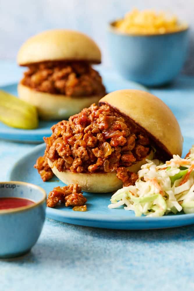 A ground chicken sloppy joes sandwich on a blue plate with coleslaw with another sandwich in the background and cheese in a bowl.