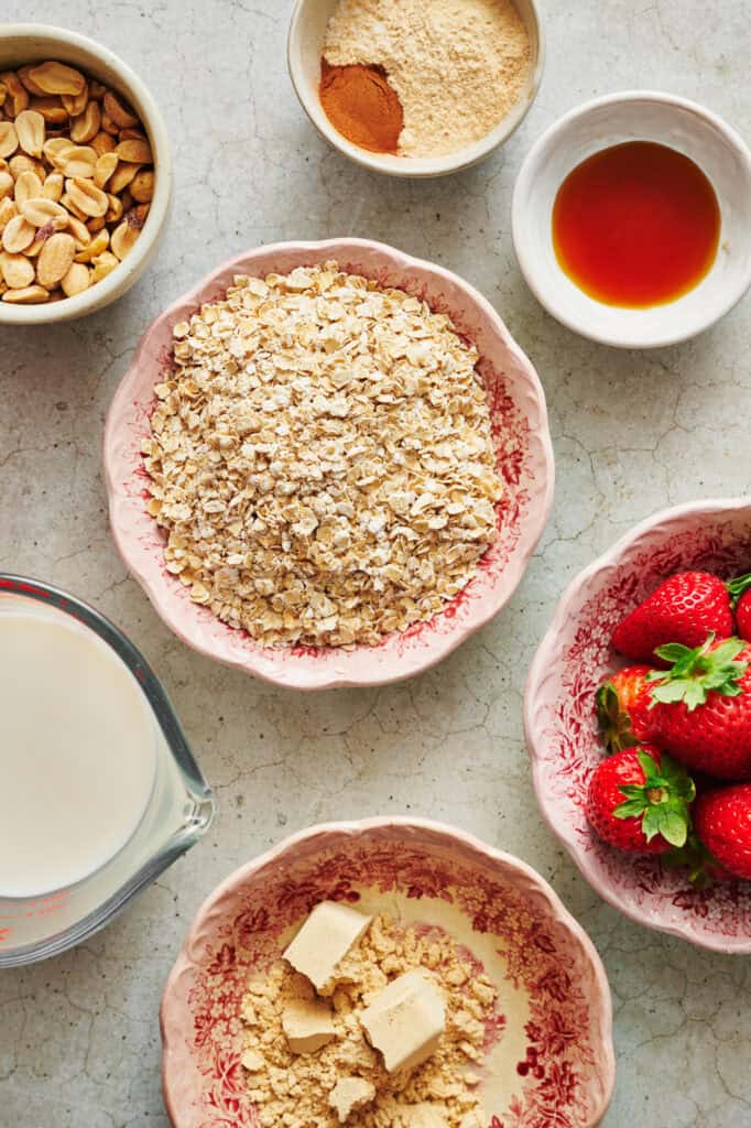 ingredients for overnight oats on a marble surface. includes oats, vanilla, protein powder, peanut butter powder, cinnamon, non-dairy milk and peanuts.