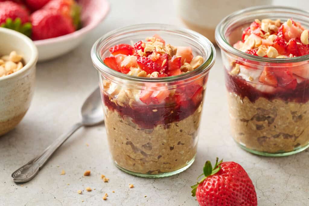 two glass jars filled with overnight oats, jelly, yogurt, chopped fruit and peanuts with a spoon on the side