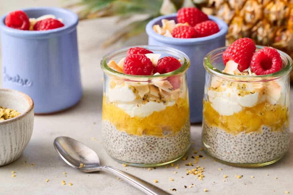 Two glass jars with pineapple chia seed pudding topped with coconut and raspberries and a spoon on the side.