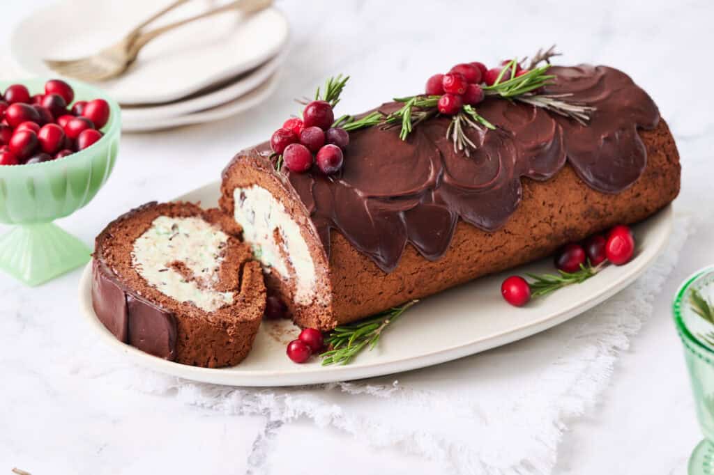 An ice cream yule log cake on a platter filled with mint chocolate chip ice cream and topped with chocolate ganache and decorated with cranberries and rosemary to look like holly and evergreen branches