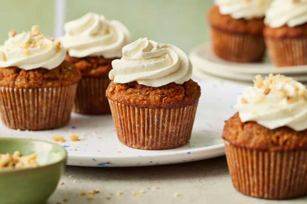 carrot cake cupcakes arranged in a scene with a green background, topped with white cream cheese frosting