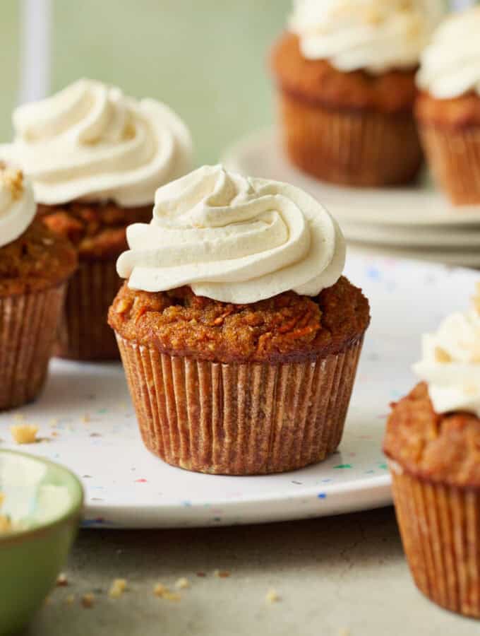 carrot cake cupcakes arranged in a scene with a green background, topped with white cream cheese frosting