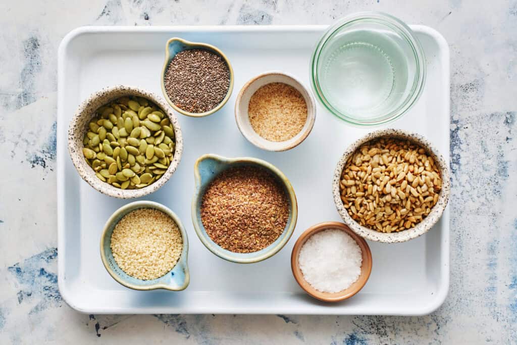 overhead view of a white sheet pan with the ingredients for flax seed crackers. Includes pumpkin seeds, flax seeds, sesame seeds, chia seeds, psyllium husk, sunflower seeds, water and salt.