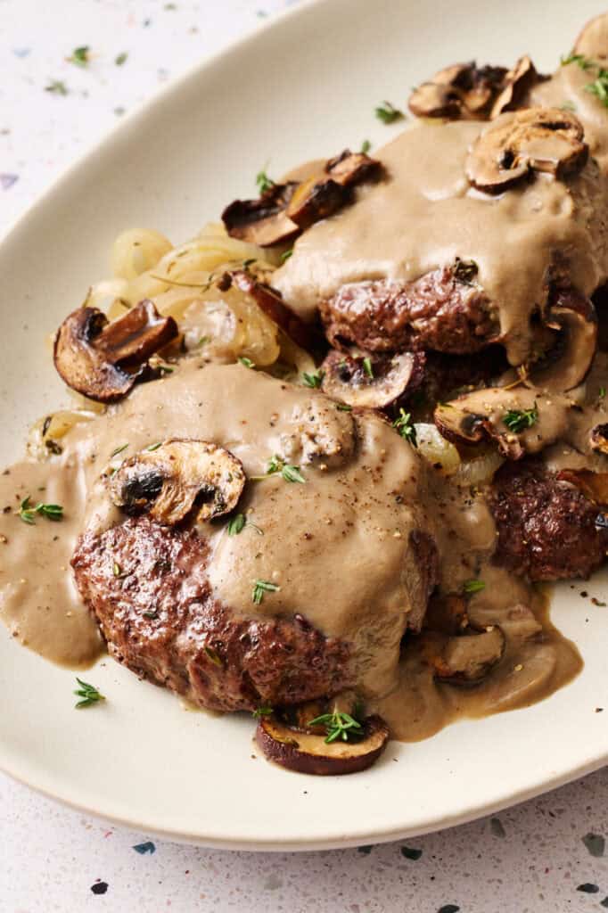 A platter with Oven Hamburger Steaks smothered in mushroom gravy with onions and mushrooms scattered around.