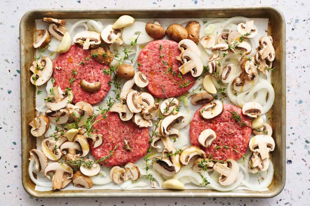 All the sheet pan components ready to be cooked, including beef patties, mushrooms, onions, thyme, salt, pepper and avocado oil