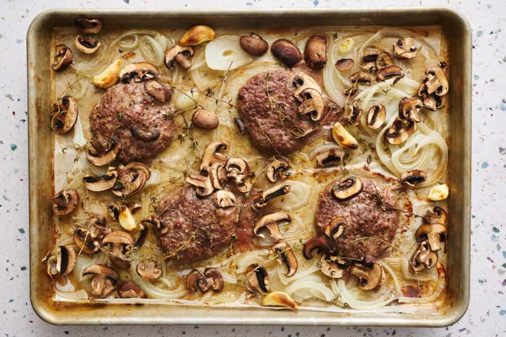 the sheet pan after roasting is done showing cooked burgers, onions, mushrooms and garlic