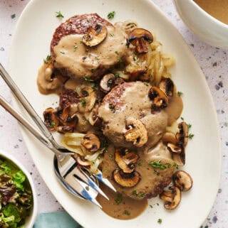 Overhead view of a platter with Oven Hamburger Steaks smothered in mushroom gravy with onions and mushrooms scattered around.