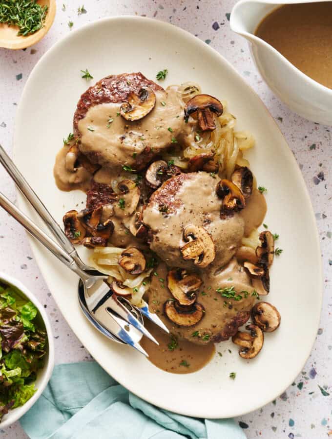 Overhead view of a platter with Oven Hamburger Steaks smothered in mushroom gravy with onions and mushrooms scattered around.