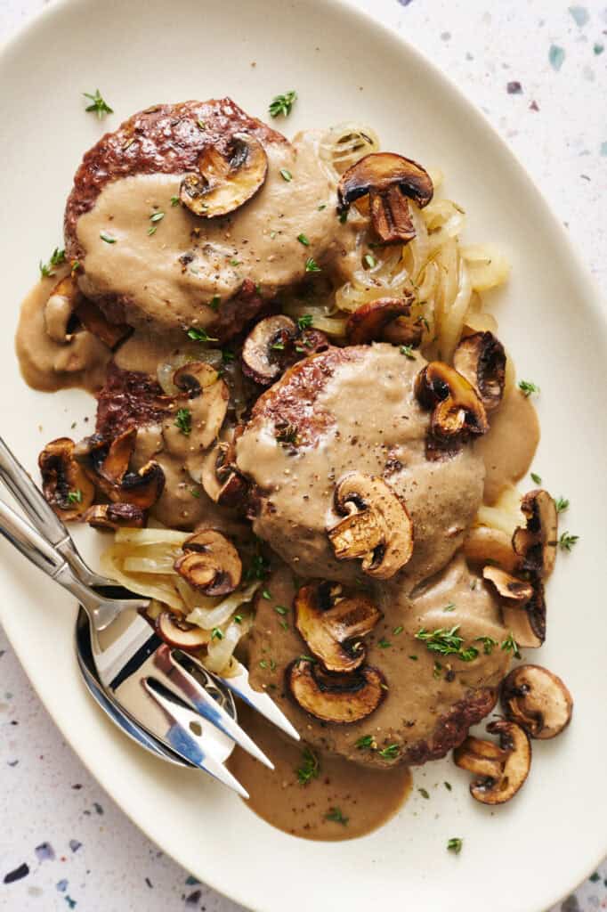 Overhead view of a platter with Oven Hamburger Steaks smothered in mushroom gravy with onions and mushrooms scattered around.