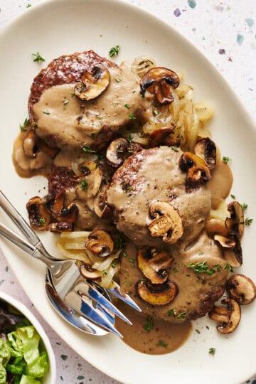 Overhead view of a platter with Oven Hamburger Steaks smothered in mushroom gravy with onions and mushrooms scattered around.