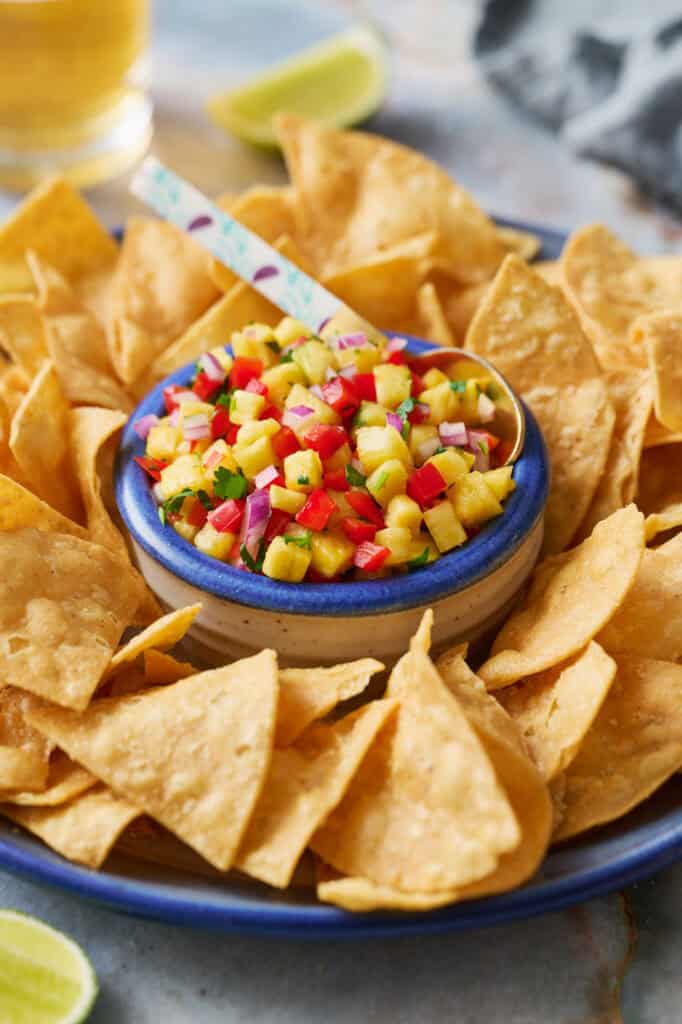 side angle view of a platter of tortilla chips with pineapple pico de gallo in the center with a spoon. beers and limes in the peripheral view.