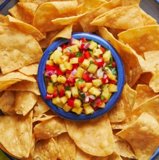 Overhead view of a platter of tortilla chips with pineapple pico de gallo in the center with a spoon. Beers and limes in the peripheral view.