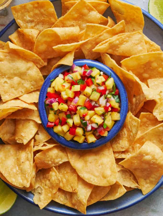 Overhead view of a platter of tortilla chips with pineapple pico de gallo in the center with a spoon. Beers and limes in the peripheral view.