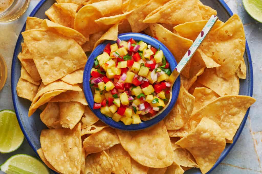 Overhead view of a platter of tortilla chips with pineapple pico de gallo in the center with a spoon. Beers and limes in the peripheral view.