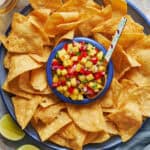 Overhead view of a platter of tortilla chips with pineapple pico de gallo in the center with a spoon. Beers and limes in the peripheral view.