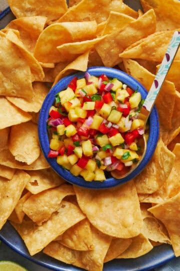 Overhead view of a platter of tortilla chips with pineapple pico de gallo in the center with a spoon. Beers and limes in the peripheral view.