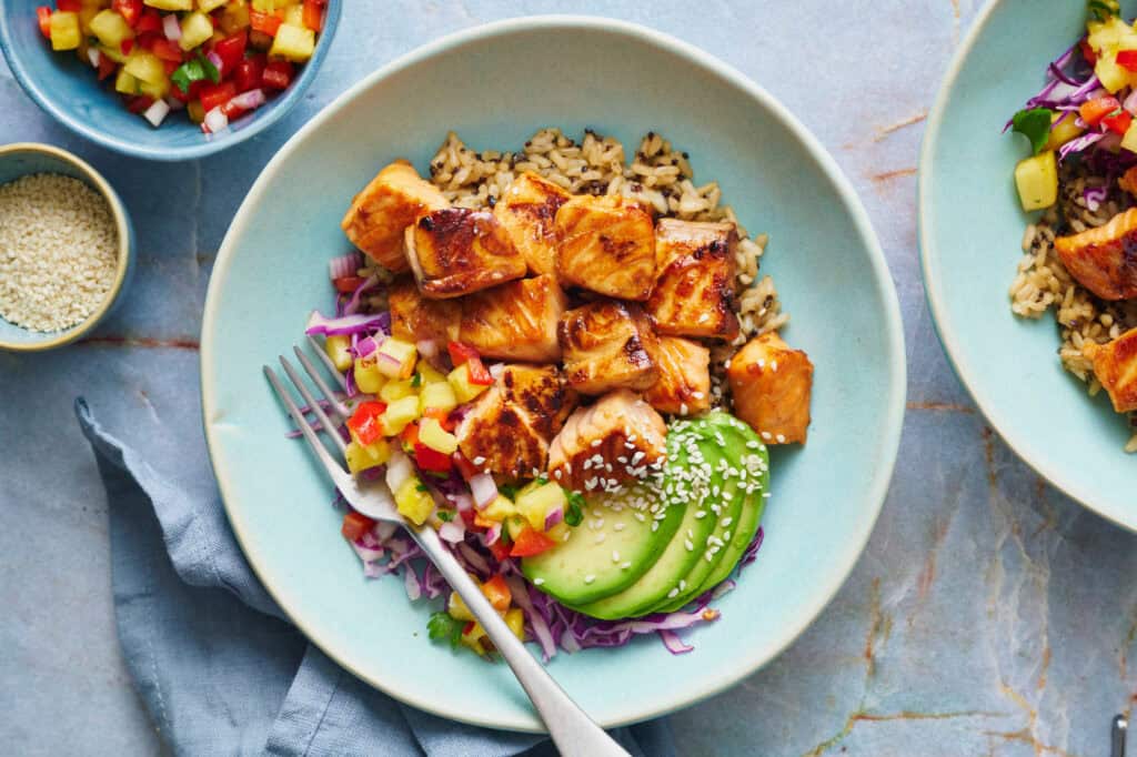 overhead of teriyaki salmon bites in a bowl with quinoa rice, avocado, salsa and cabbage with sesame seeds on the the side