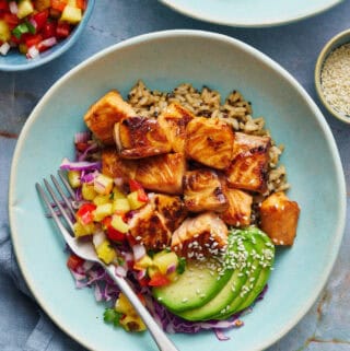 overhead of teriyaki salmon bites in a bowl with quinoa rice, avocado, salsa and cabbage with sesame seeds on the the side