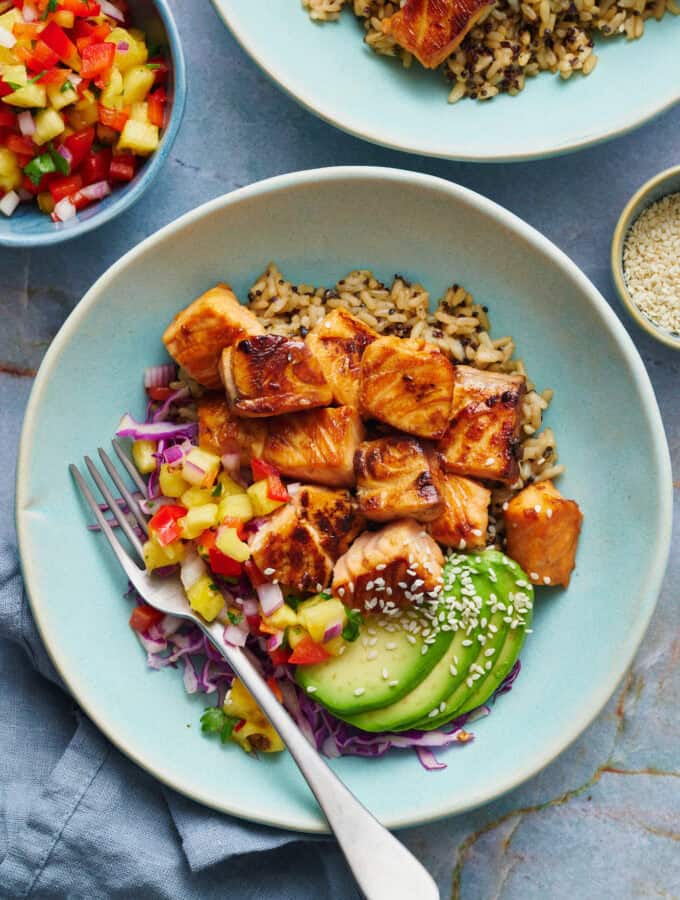 overhead of teriyaki salmon bites in a bowl with quinoa rice, avocado, salsa and cabbage with sesame seeds on the the side