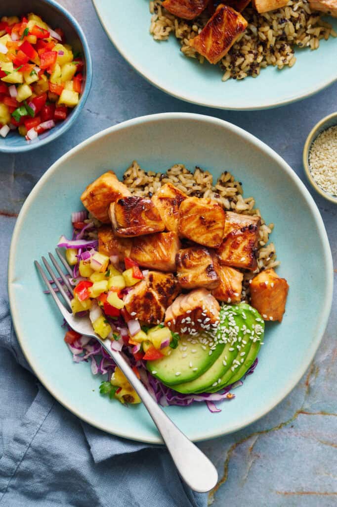 overhead of teriyaki salmon bites in a bowl with quinoa rice, avocado, salsa and cabbage with sesame seeds on the the side