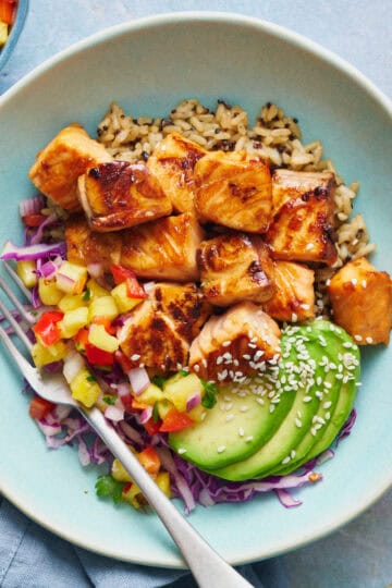 overhead of teriyaki salmon bites in a bowl with quinoa rice, avocado, salsa and cabbage with sesame seeds on the the side