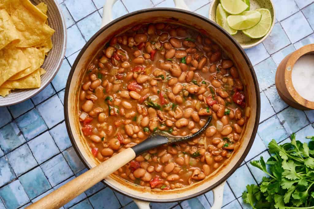 a white pot filled with vegan charro beans with a ladle scooping out some beans with tomato and spices