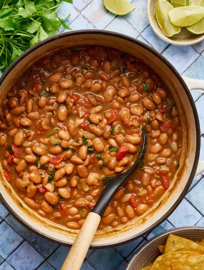 a white pot filled with vegan charro beans with a ladle scooping out some beans with tomato and spices