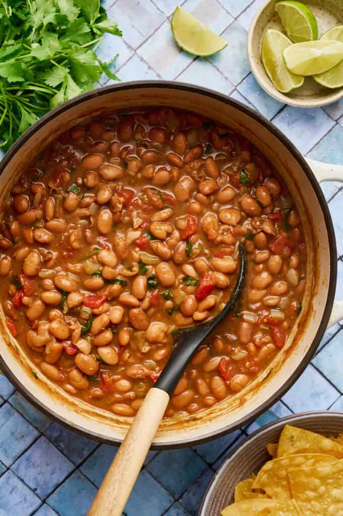 a white pot filled with vegan charro beans with a ladle scooping out some beans with tomato and spices