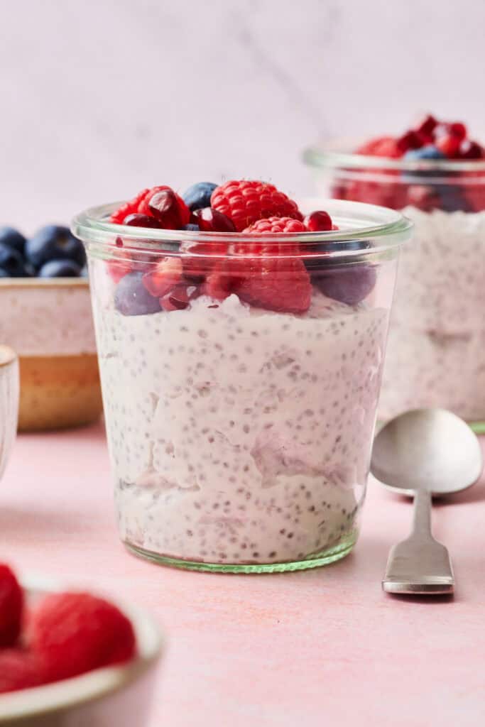 chia yogurt parfaits on a pink background with fresh raspberries, blueberries and pomegranate seeds surrounding