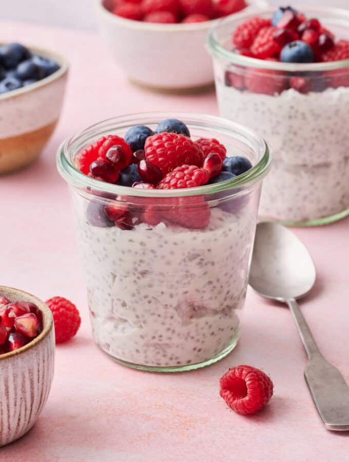 chia yogurt parfaits on a pink background with fresh raspberries, blueberries and pomegranate seeds surrounding