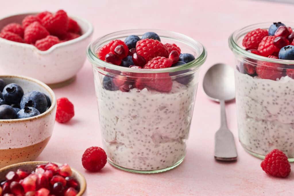 chia yogurt parfaits on a pink background with fresh raspberries, blueberries and pomegranate seeds surrounding