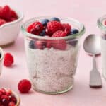 chia yogurt parfaits on a pink background with fresh raspberries, blueberries and pomegranate seeds surrounding