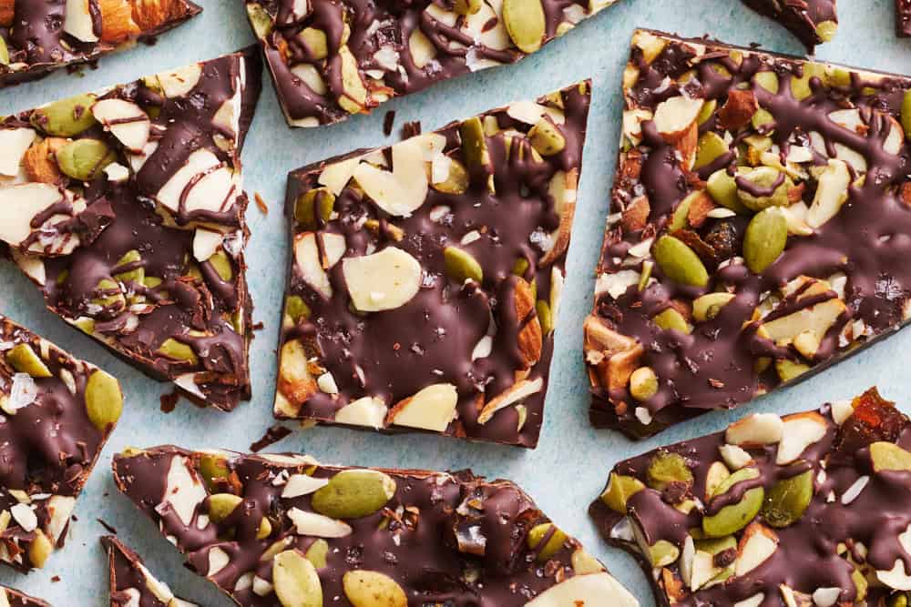 overhead view of chocolate bark with sea salt, almonds, pumpkin seeds and dates cut up on a blue surface