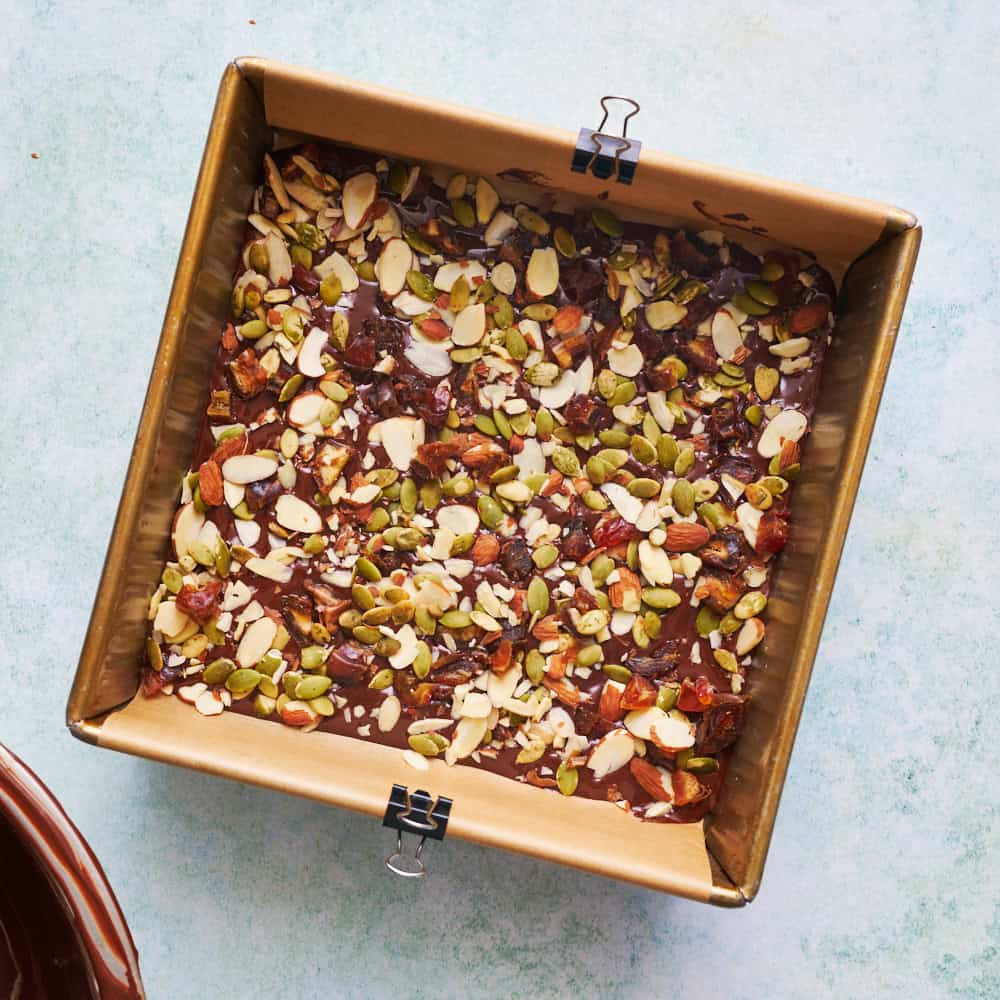 overhead view of chocolate bark with sea chocolate, salt, almonds, pumpkin seeds and dates in an 8 inch pan being made