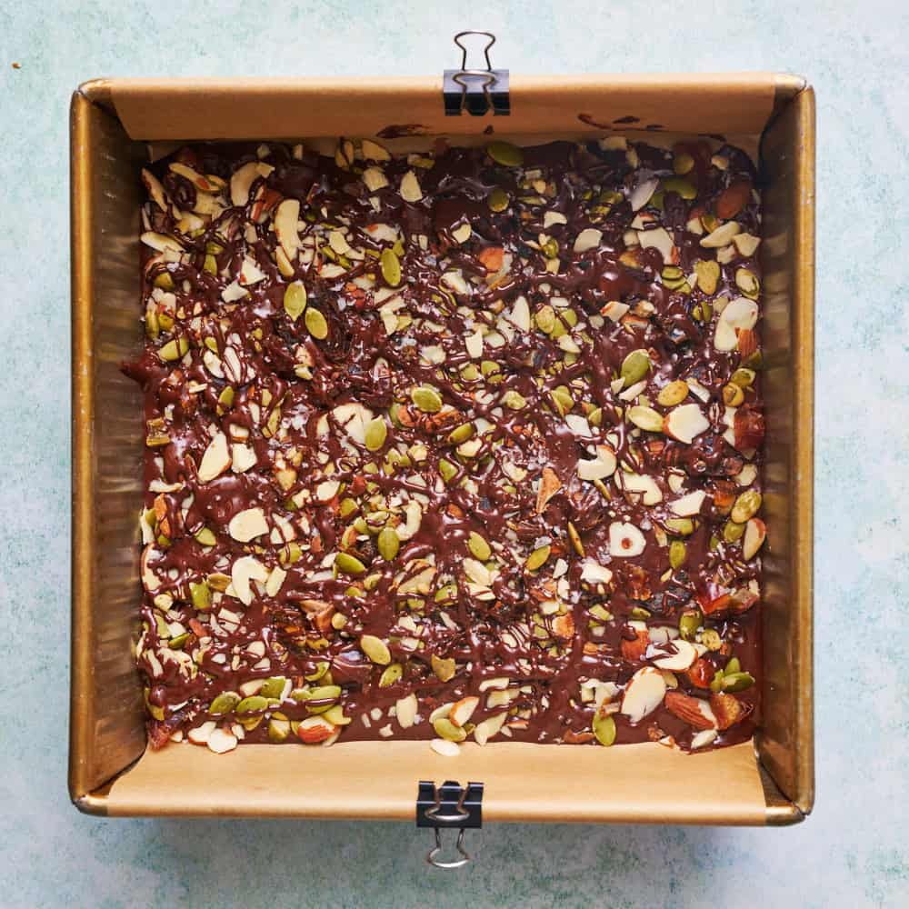 overhead view of chocolate bark with sea salt, almonds, pumpkin seeds and dates in an 8 inch pan after chocolate was drizzled on top