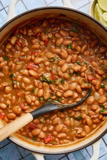 a white pot filled with vegan charro beans with a ladle scooping out some beans with tomato and spices