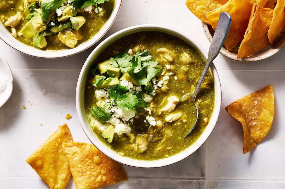 a bowl of chile verde topped with avocado, cotija cheese and cilantro with chips on the side