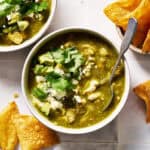 a bowl of chile verde topped with avocado, cotija cheese and cilantro with chips on the side