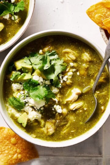 a bowl of chile verde topped with avocado, cotija cheese and cilantro with chips on the side
