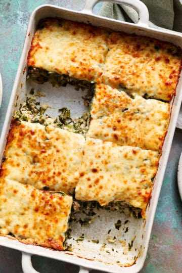 A large dish filled with a spinach, cheese and beef casserole with a piece taken out and put on a plate with a fork