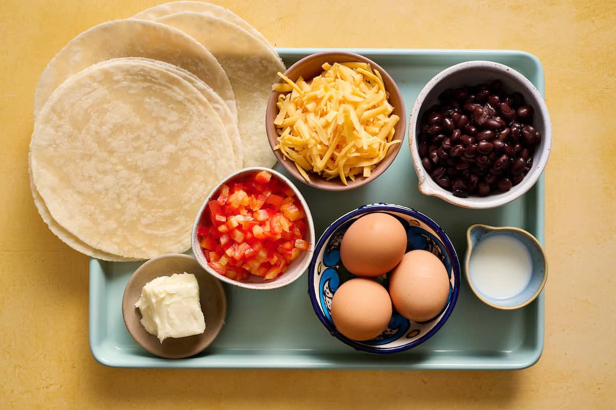 ingredients to make breakfast quesadillas including butter, tortillas, eggs, milk, black beans, cheese and diced pepper