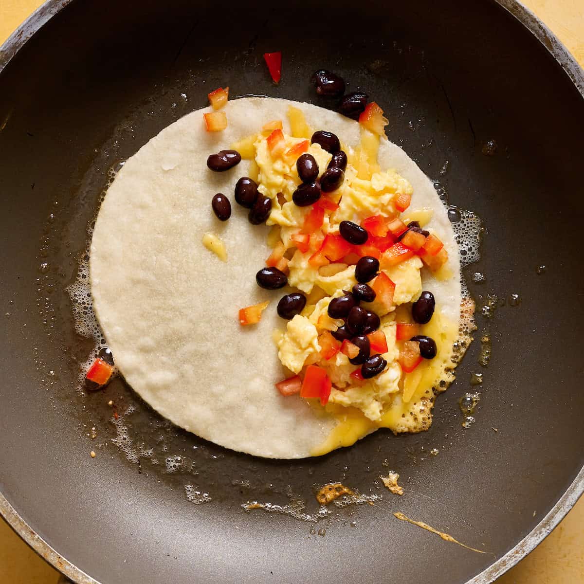 a tortilla in a skillet topped with eggs, cheese, peppers and beans on one half